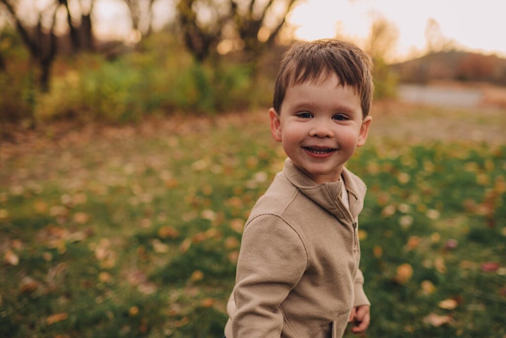 A little boy smiles at the camera while running in a beige sweatshirt and jeans.