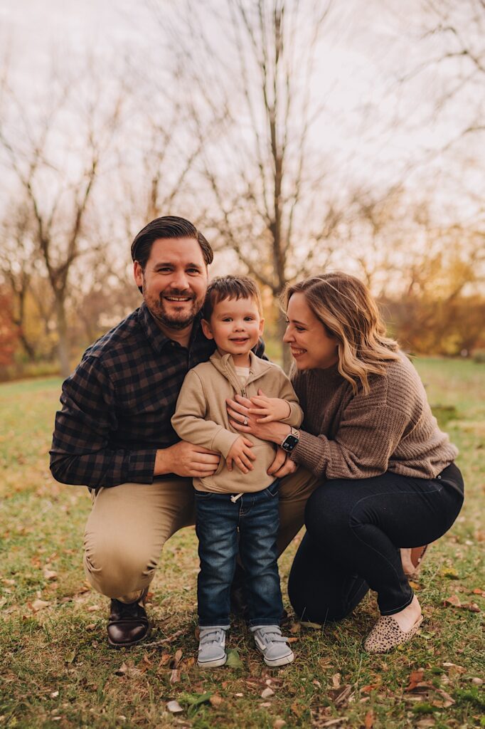 A family sits together smiling at their little boy in a forest preserve in the fall