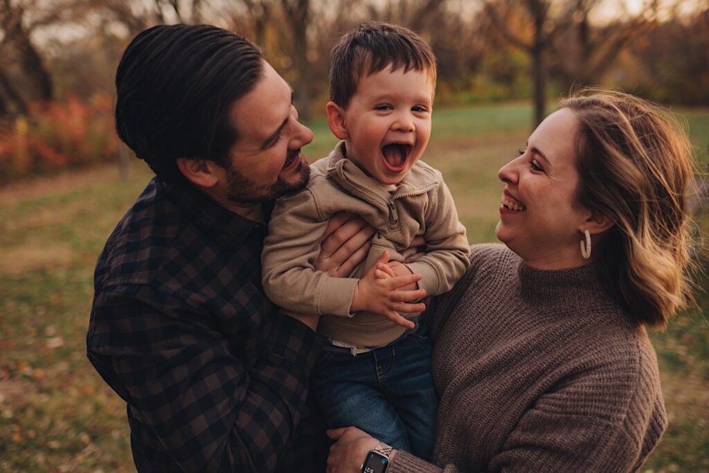 A couple holds their little boy between them while making him laugh while playing at a forest preserve in wheaton.